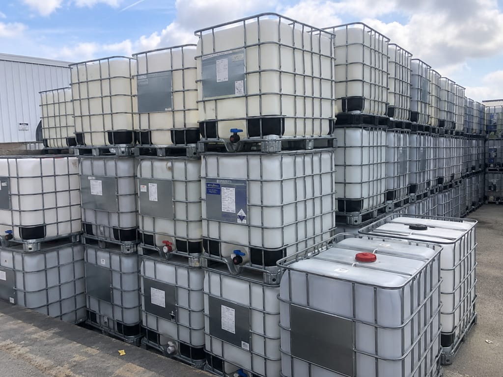 Rows of used 275 gallon composite IBC totes stacked two high in an outdoor yard under a blue sky