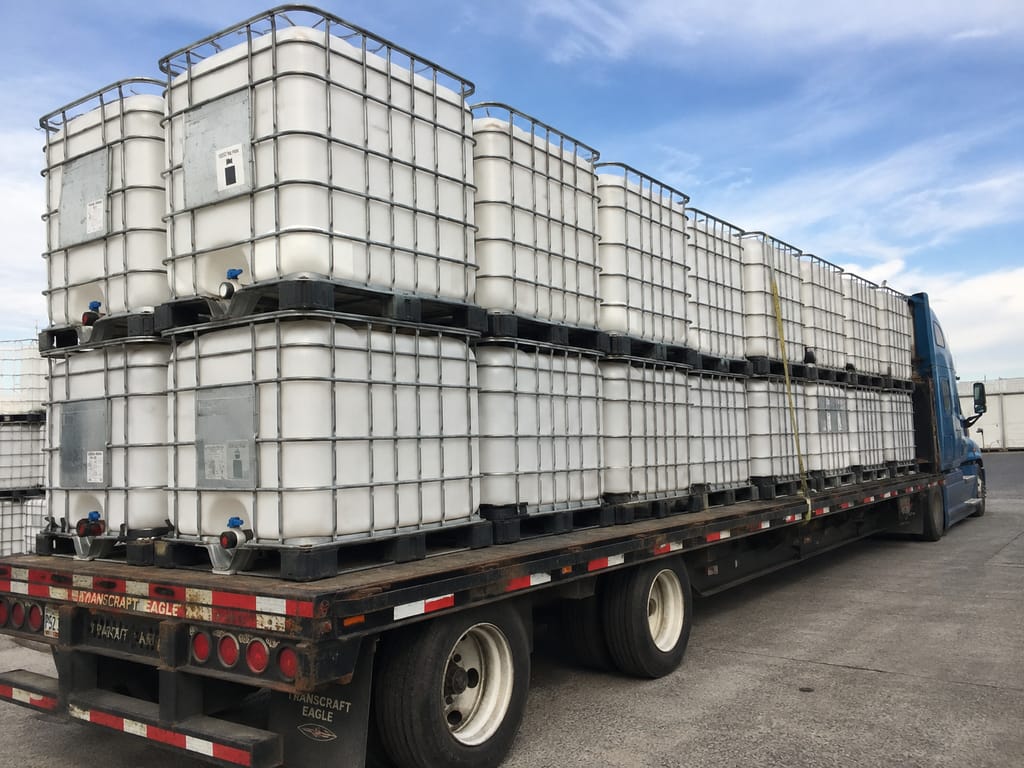 Flatbed trailer loaded with two rows of IBC totes secured with straps, hooked to a blue tractor unit